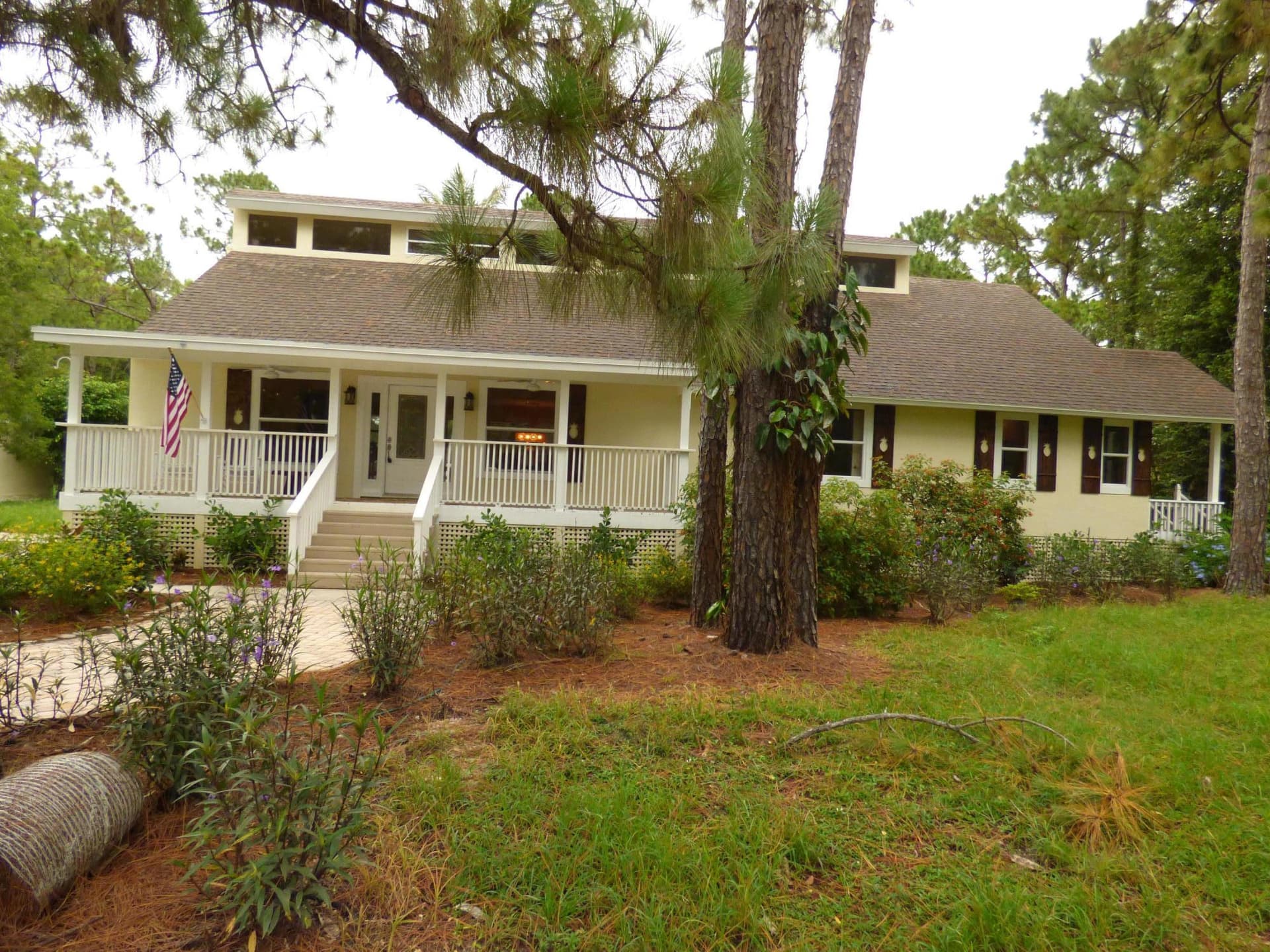 Cream ranch home with a wraparound porch and dark walnut pineapple-cutout shutters, framed by tall pines