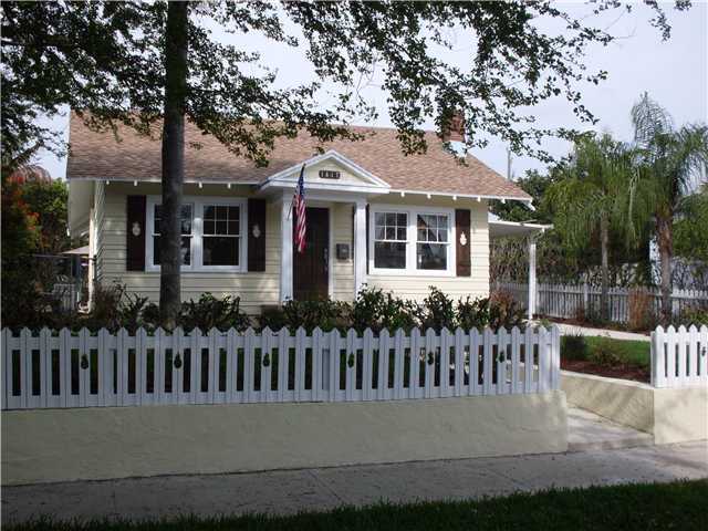 Pale yellow cottage bungalow with dark walnut pineapple-cutout shutters behind a white picket fence